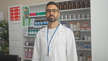 A confident hispanic man stands in a pharmacy, wearing a lab coat and glasses.