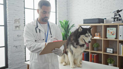 A young hispanic man in a white coat writes on a clipboard while a husky dog observes in a veterinarian clinic.