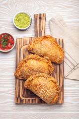Three freshly baked empanadas with sesame seeds on a wooden board, white background, served with tomato salsa and guacamole. Traditional dish of Spain and Latin America