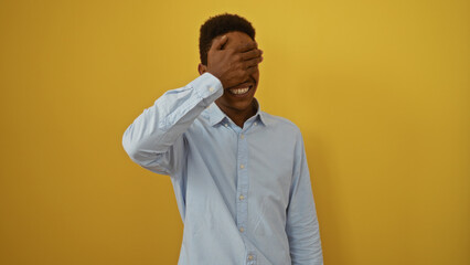Fototapeta premium Young man in blue shirt covering eyes with hand against isolated yellow background, smiling playfully.