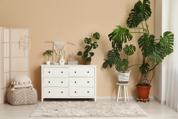 Monstera houseplants, chest of drawers, pouf with cushion and folding screen near beige wall