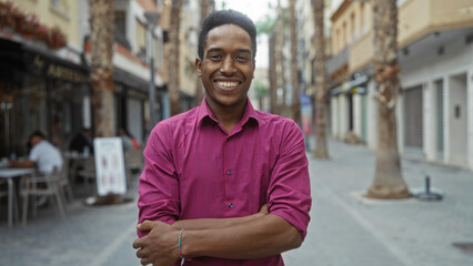 Young man with crossed arms smiles confidently on an urban street, showcasing a casual atmosphere with people and cafes in the background.