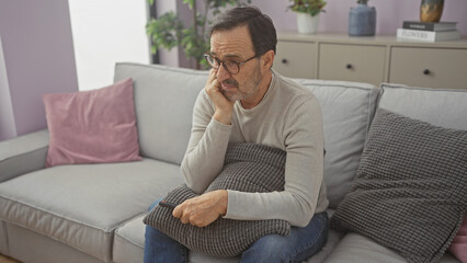 Pensive hispanic man sitting on couch at home, holding pillow, wearing glasses, casual, alone,...