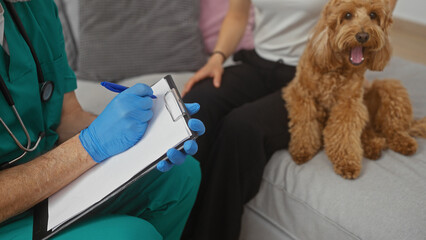 A veterinarian in scrubs takes notes beside a seated man and his attentive poodle in a bright living room.