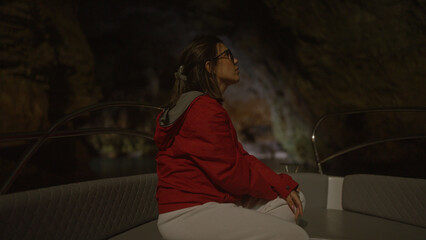 Obraz premium A young hispanic woman in a red jacket sits thoughtfully on a boat in a dimly lit cove in polignano a mare, puglia, italy, with the cave's rocky walls enclosing the serene sea around her.