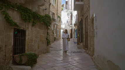 Obraz premium A young hispanic woman strolls through the charming stone streets of polignano a mare in puglia, italy, basking in the warm european ambiance.