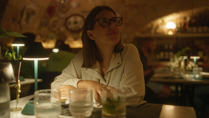 A young, beautiful hispanic woman wearing glasses sits at a table in an indoor restaurant in italy, enjoying a cozy evening surrounded by soft lighting and elegant decor.