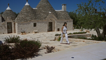 A young, beautiful hispanic woman in a white outfit walks outside historic trulli houses on a sunny...