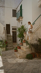 A young, beautiful hispanic woman stands on stone steps surrounded by potted plants in the charming old town streets of locorotondo, puglia, italy.