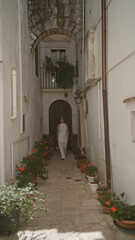 A beautiful young hispanic woman walks down a narrow street in the charming old town of locorotondo, puglia, italy, surrounded by white buildings and vibrant potted plants.