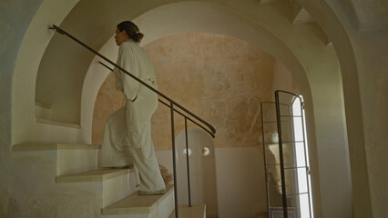A beautiful young hispanic woman ascends an indoor staircase in a typical italian house, showcasing the architectural charm of a rustic masseria.