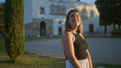 A beautiful young hispanic woman stands outdoors at a villa in puglia, italy, with the sunlight highlighting her face and the elegant house in the background.