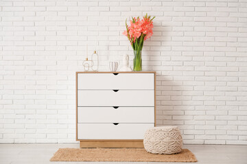 Vase with gladiolus flowers, Moai statue and candles on commode near white brick wall in room