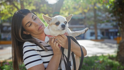 A young hispanic woman holds a chihuahua dog in an urban park setting, conveying companionship and leisure outdoors.