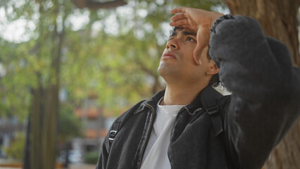 A young, handsome hispanic man shields his eyes while looking upward in an urban park setting.