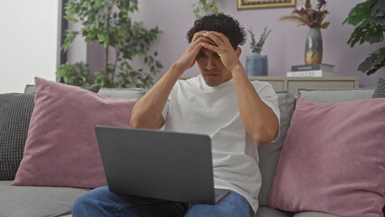 Stressed hispanic man with laptop sitting on couch in a modern apartment living room, exhibiting...
