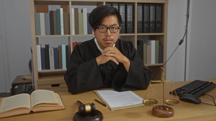 Handsome young asian man dressed as a judge sits in an office with legal books, a gavel, and scales...