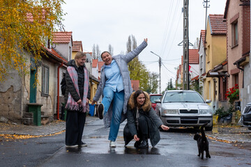 Obraz premium Three happy women enjoying moment with black cat on picturesque street lined with colorful houses and autumn trees. Concept of companionship, charm of small towns, and enjoying simple outdoor moments.