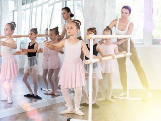 Fototapeta premium Adult woman teacher helping group of girls dancers practicing plie movement at barre in studio
