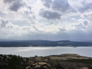 Stausee inmitten einer kargen Landschaft, umgeben von felsigen Hügeln. Ein beeindruckender Wolkenhimmel dominiert die Szenerie und verleiht ihr Dramatik