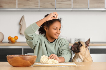 Cute African-American girl with raw dough and French bulldog at table in kitchen
