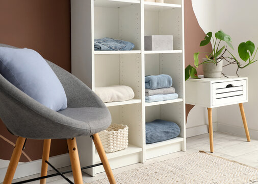Shelving unit with clothes, stylish armchair and houseplant near color wall