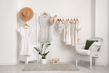 Rack with stylish female clothes, hats, wicker armchair and houseplant near light wall in room