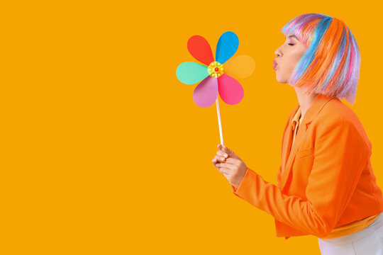 Beautiful young happy woman in colorful wig with toy windmill blowing on yellow background