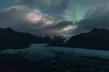 Aurora borealis glowing over glacier lagoon with mountains and dark cloudy sky in iceland