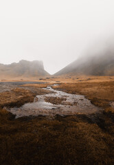 Serene stream flows through a misty valley in iceland, surrounded by majestic mountains. The tranquil atmosphere is enhanced by foggy weather, creating a calm scenery with brown and gray tones