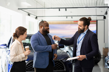 Showroom agent completes paperwork accompanying vehicle sale, shaking hands with clients and offering car keys. Salesperson in dealership signing contract with customers, reaching agreement