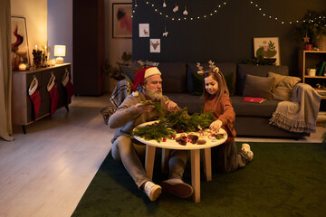 Father and daughter sitting on floor and decorating Christmas tree with ornaments, enjoying holiday spirit together in warm, festive living room with stockings hanging on wall