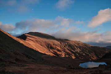 Serene mountain landscape at sunrise. Golden hour light illuminates the rugged peaks and a tranquil lake nestled below.  A sliver of moon hangs in the clear blue sky.