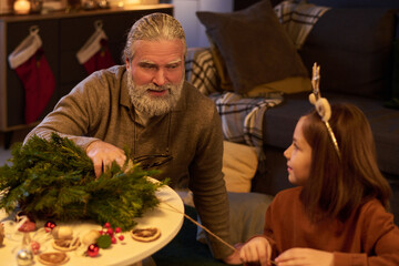 Elderly man engaging with young girl while making holiday decorations, creating festive atmosphere in living room. Christmas stockings and decorations visible in background