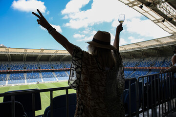 Fototapeta premium A woman with a hat raises her hands to the sides against the background of a football stadium and a cloudy blue sky
