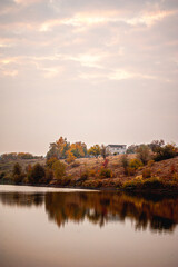Hills near a lake with orange grass, trees and bushes, on which a two-story brick house is located, against the background of a bright dawn sky with clouds