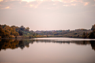 a calm lake, with reflections, against the backdrop of banks with reeds and a bright dawn sky