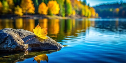 Fototapeta premium A Serene Composition of a Yellow Leaf on a Gray Rock Surrounded by Tranquil Blue Water and Blurred Green Trees Under a Clear Sky for Nature Photography