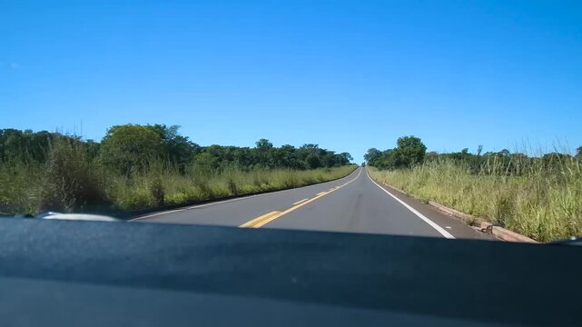 Driving the car on a Brazilian highway surrounded by rural areas and forest on a blue sky day.