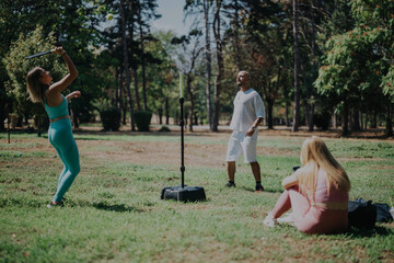 Three friends relax and play games in a scenic park setting, enjoying a casual day outdoors. A woman hits a ball with a racket while others watch, surrounded by greenery and open space.