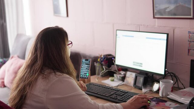 Woman sitting at a desk, working from home on multiple computer screens, focusing on tasks with concentration and efficiency