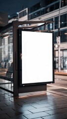 A mockup of a blank white vertical digital billboard poster at a city street bus stop sign at night. The billboard is illuminated.	