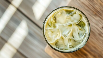 A top view of a refreshing iced matcha latte with a beautiful green swirl of matcha and milk, served in a glass over ice. The drink sits on a minimalist wooden table