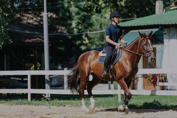 A man is practicing horseback riding at an outdoor equestrian arena. The rider is focused, wearing appropriate safety gear while the horse moves gracefully in a fenced training area.
