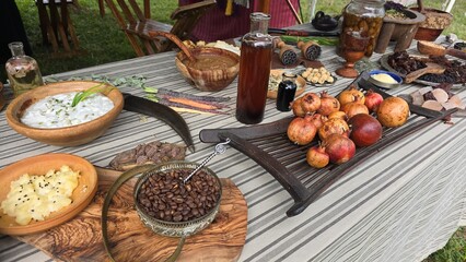 Table with rustic dishes, including pomegranates, coffee beans, creamy dishes, dried fruits, and bottles of liquids. The setting suggests a historical or cultural culinary display.