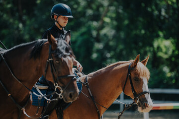 A young equestrian rides two horses in a sunny outdoor arena, exuding focus and confidence. The lush green background highlights the beauty and grace of the equestrian sport.