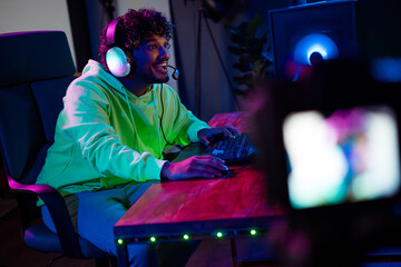 Photo of handsome young man video recording camera sit table play gamer computer game late night blue purple indoors house late neon light © deagreez