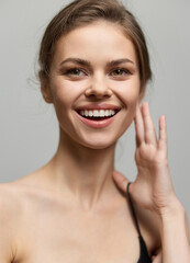 Fototapeta premium Cheerful young woman smiling and touching her face with one hand, showcasing confidence and joy. Beauty and self-care concept against a soft neutral background.