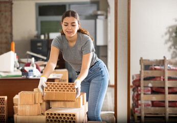 Interested young attractive woman in gray t-shirt and jeans working on her house renovation, preparing red bricks for masonry