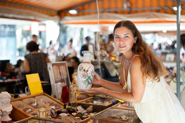 Girl in flea market store look at Chinese porcelain painted vase, antique gifts. Female buyer holds...